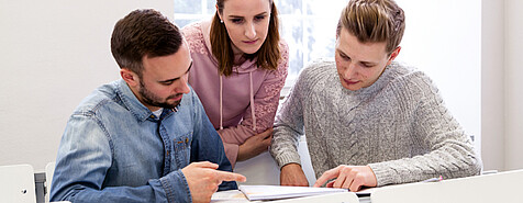 Drei Personen diskutieren in der Universität Hohenheim, eine stehend und zwei sitzend, über die auf einem Tisch liegenden Unterlagen zu den Master- und Bachelor-Studiengängen.